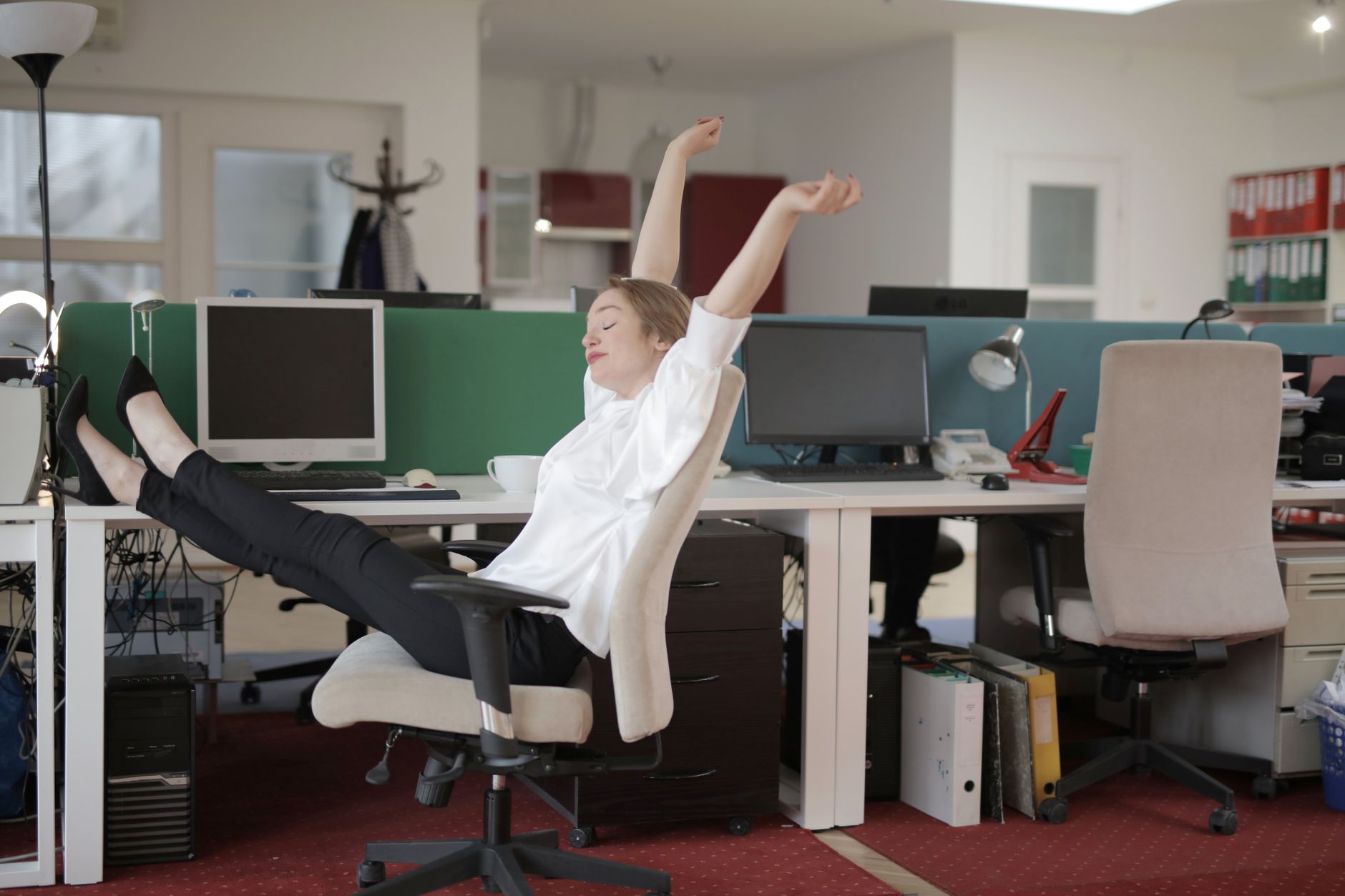 Employée souriante assise, jambes croisées sur bureau, s’étirant au travail.