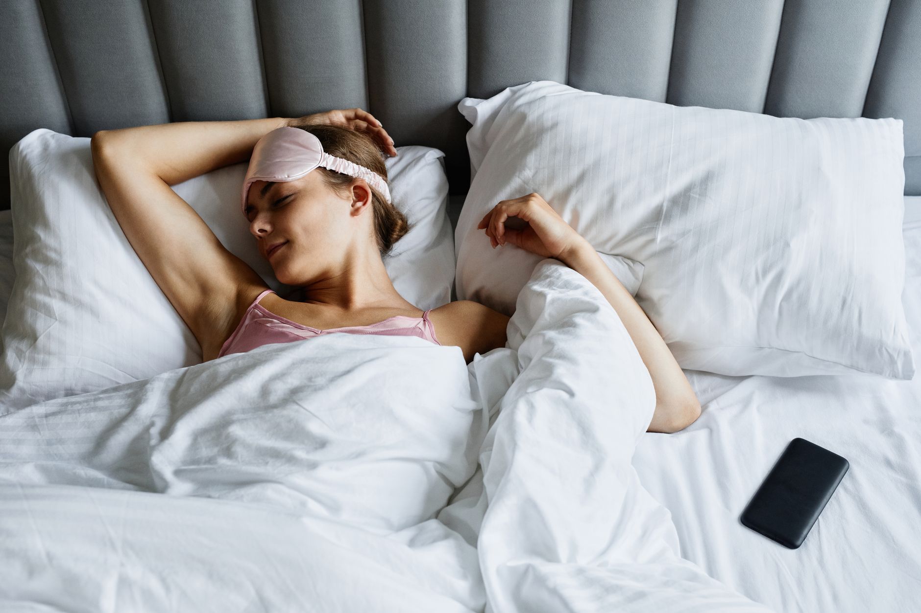 Femme reposant au lit avec un masque pour les yeux et un smartphone à côté.