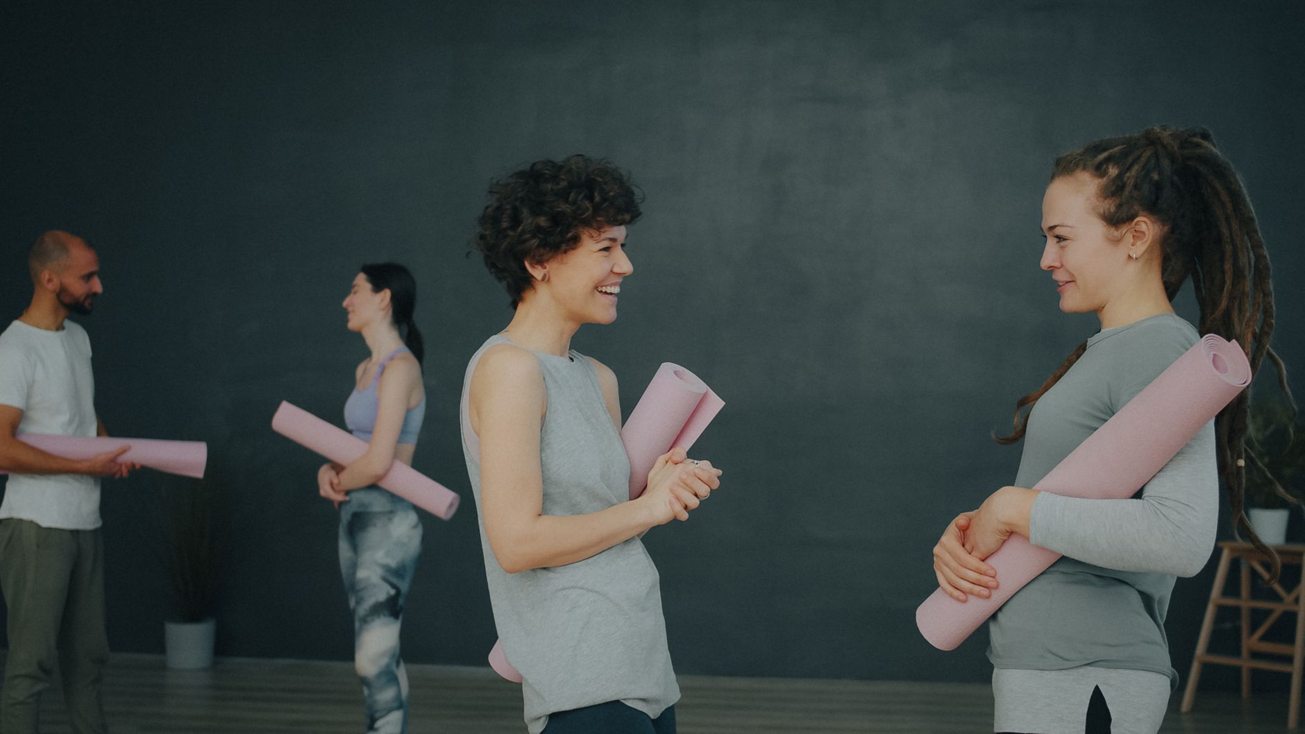 Quatre adultes en cours de yoga intérieur, discutant avec tapis en main.
