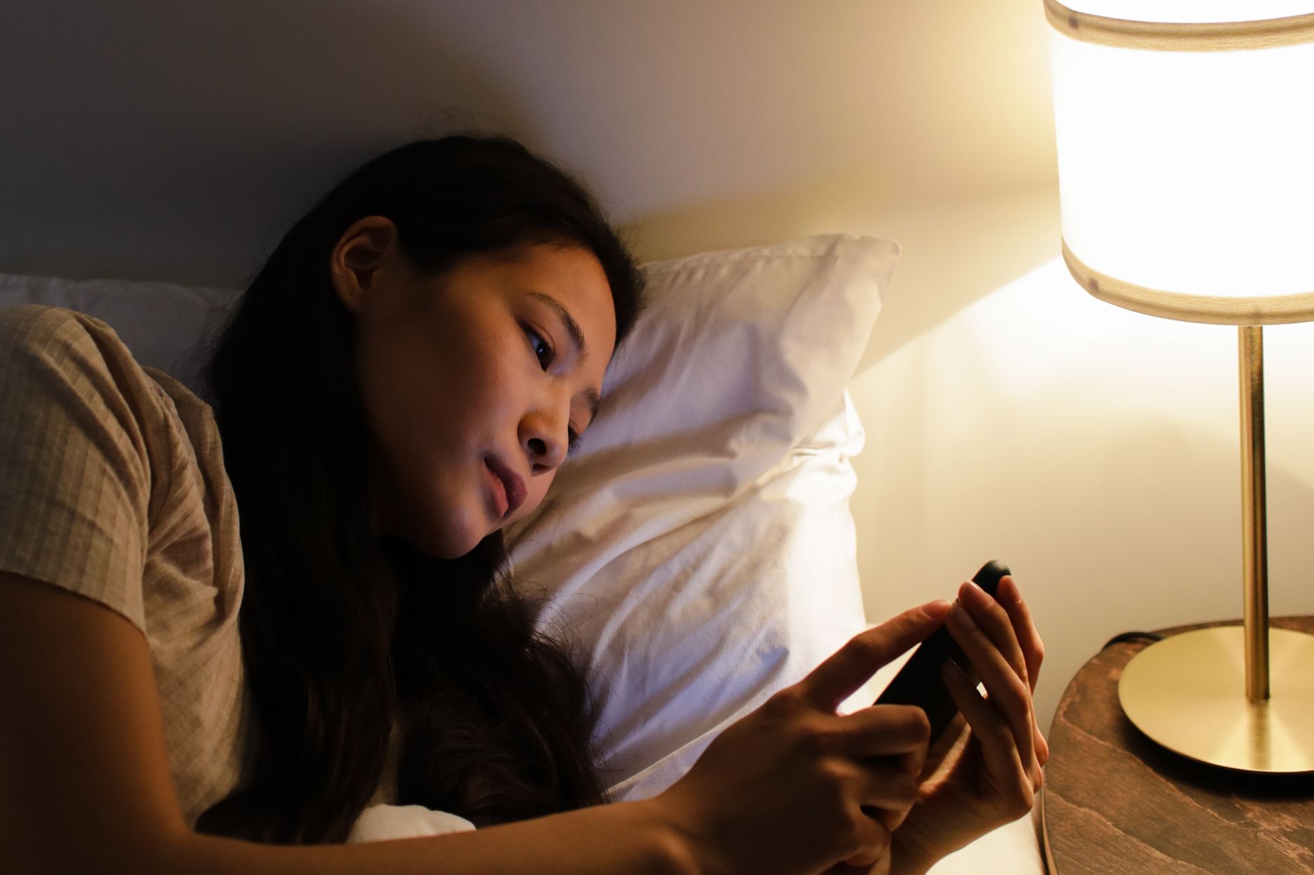 jeune femme allongée dans son lit, la nuit, qui lit un message sur son téléphone à la lumière d'une lampe de chevet