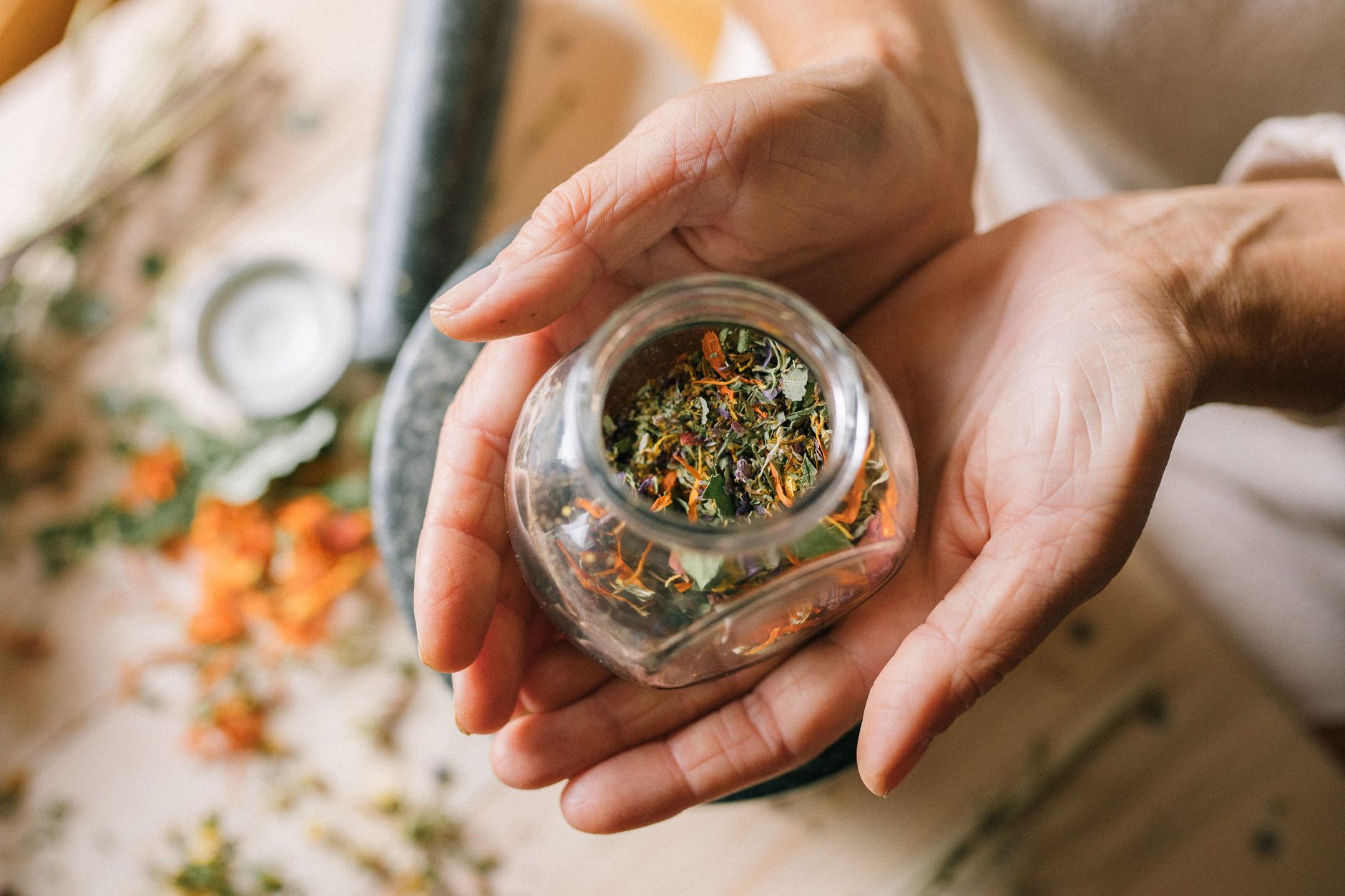 mains qui tiennent un bocal en verre rempli de plantes séchées au-dessus d'une table de préparation